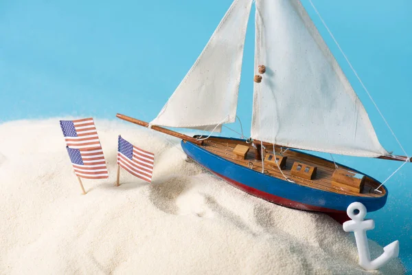 American national flags in white sand near miniature ship and anchor isolated on blue — Stock Photo