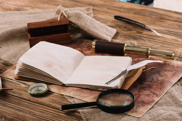 Libro abierto con páginas en blanco cerca del telescopio, brújula y lupa en la mesa de madera con hessian y mapa del mundo - foto de stock