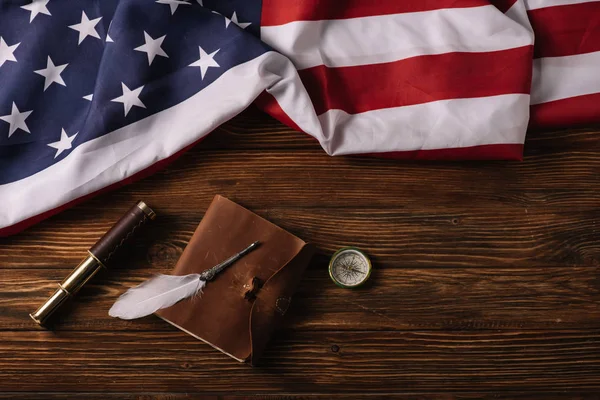 Vista superior do caderno de couro, telescópio, nib e bússola na superfície de madeira com bandeira nacional americana — Fotografia de Stock