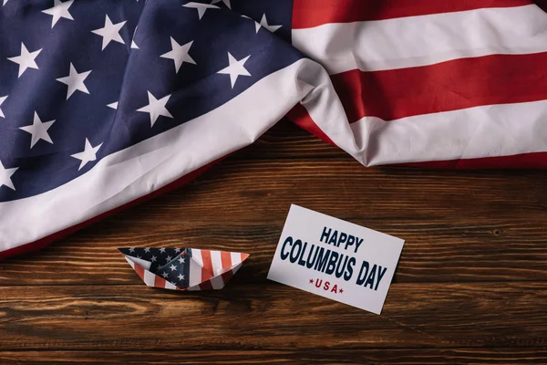 Top view of card with happy Columbus Day inscription near paper boat on wooden surface with American national flag — Stock Photo