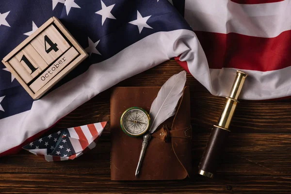 Vista superior del calendario de madera con fecha 14 de octubre, barco de papel, plumín, brújula, telescopio y cuaderno de cuero en la superficie de madera con bandera nacional estadounidense - foto de stock