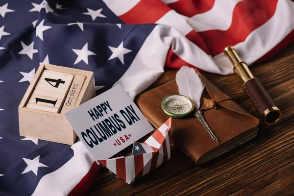Card with happy Columbus Day inscription near calendar, paper boat, nib, compass, telescope and notebook on wooden surface with American national flag — Stock Photo