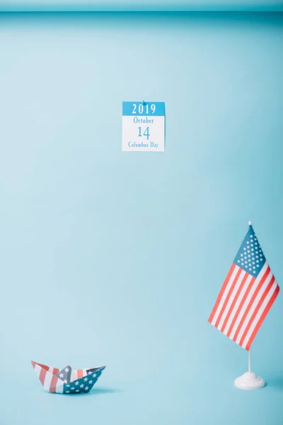 Calendário de papel com inscrição do Dia de Colombo perto de barco de papel e bandeira nacional americana em fundo azul — Fotografia de Stock