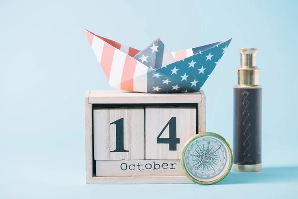 Barco de papel con patrón de bandera estadounidense en calendario de madera con fecha 14 de octubre cerca de brújula y telescopio sobre fondo azul - foto de stock