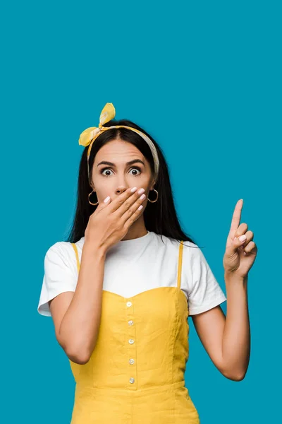 Потрясенная девушка, прикрывающая рот, указывая пальцем, изолированным на синий — стоковое фото