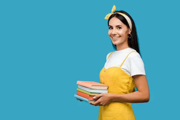 Mujer feliz en diadema sosteniendo libros y mirando a la cámara aislada en azul - foto de stock