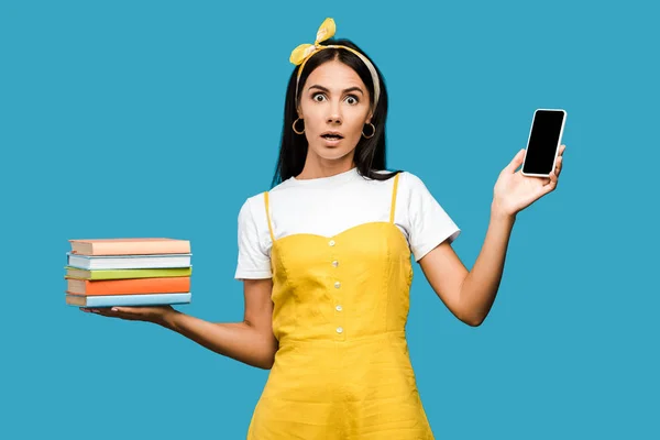 Schockierte Frau hält Bücher und Smartphone mit leerem Bildschirm isoliert auf blauem Grund — Stockfoto