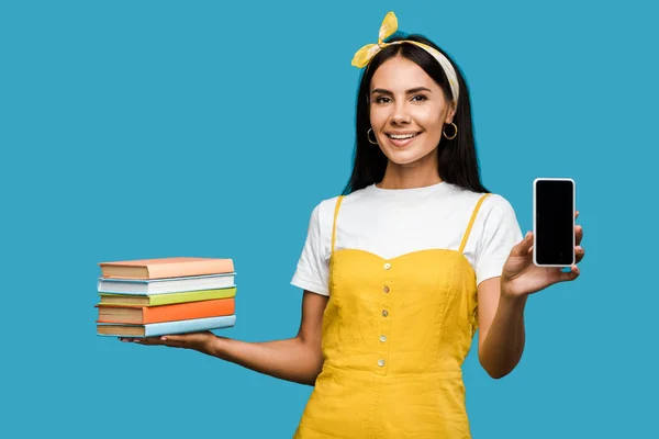 Positive woman holding books and smartphone with blank screen isolated on blue — Stock Photo