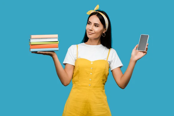 Молодая женщина держит книги и смартфон с чистым экраном изолированы на синий — стоковое фото