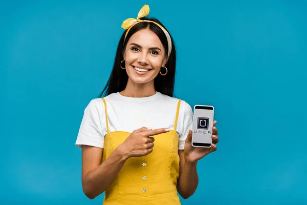 KYIV, UCRANIA - 8 de agosto de 2019: chica feliz señalando con el dedo al teléfono inteligente con la aplicación uber en la pantalla aislada en azul - foto de stock