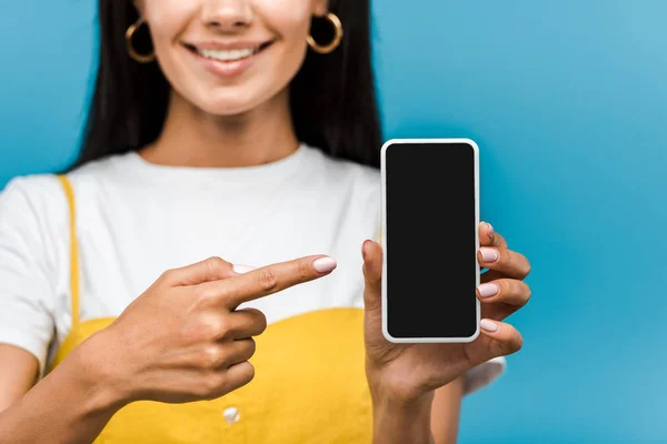 Обрезанный вид девушки, указывающей пальцем на смартфон с пустым экраном, изолированным на синий — стоковое фото