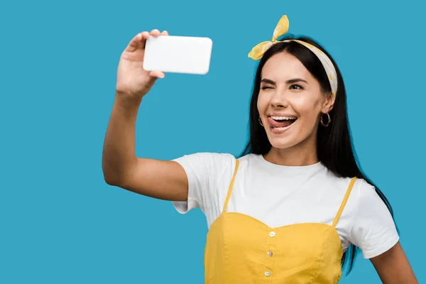 Mujer joven tomando selfie en el teléfono inteligente mientras muestra la lengua aislada en azul - foto de stock