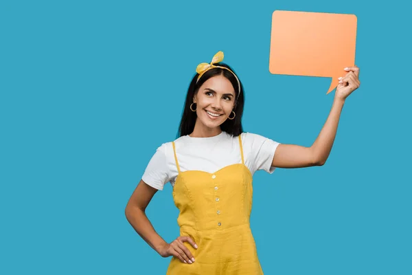 Menina feliz segurando bolha de fala laranja e de pé com a mão no quadril isolado no azul — Fotografia de Stock