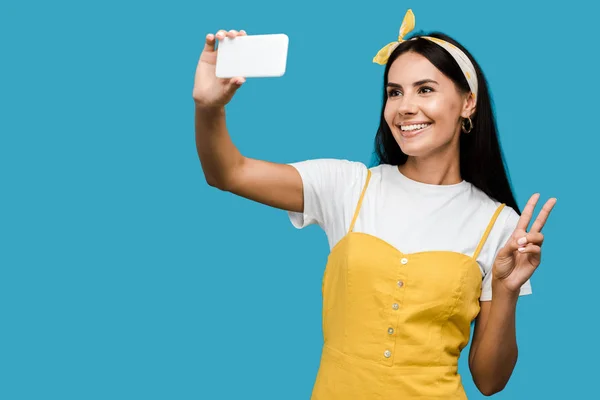 Enfoque selectivo de la mujer alegre tomando selfie en el teléfono inteligente, mientras que muestra signo de paz aislado en azul - foto de stock