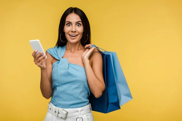 Excité jeune femme regardant la caméra et tenant smartphone avec des sacs à provisions isolés sur orange — Photo de stock