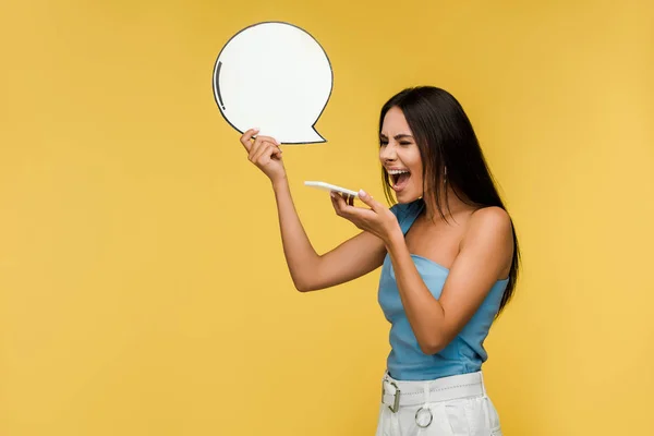 Emotional girl holding blank speech bubble and screaming while using smartphone isolated on orange — Stock Photo