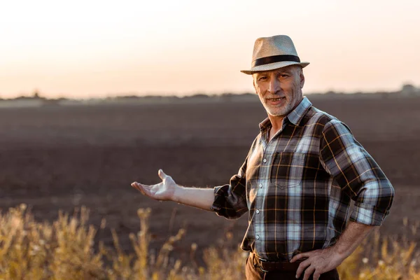 Selective focus of cheerful self-employed farmer in straw hat gesturing near wheat field — Stock Photo