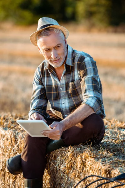 Agriculteur âgé tenant tablette numérique tout en étant assis sur la balle de foin — Photo de stock