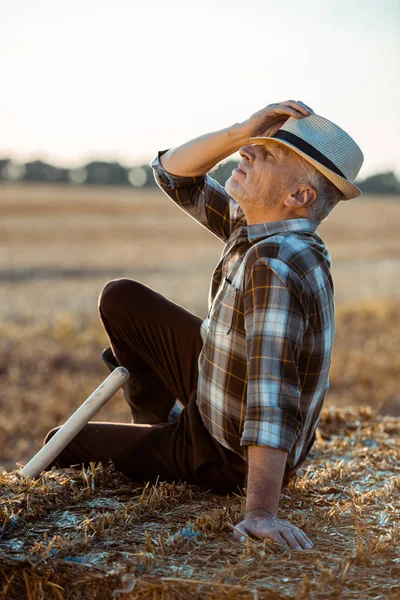 Happy bearded farmer sitting on bale of hay and touching straw hat — Stock Photo