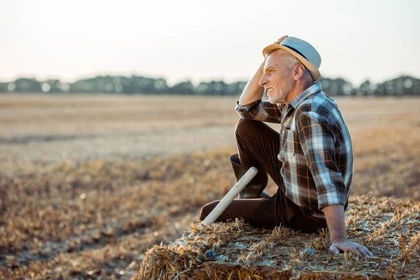 Agricultor barbudo alegre sentado no fardo de feno e tocando chapéu de palha — Fotografia de Stock