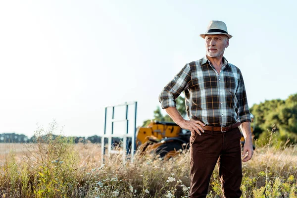 Старший чоловік у солом'яному капелюсі стоїть з рукою на стегні біля трактора — стокове фото