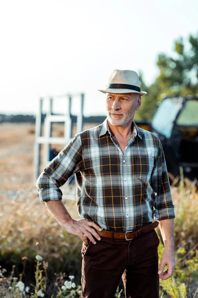 Selbstständiger Landwirt mit Strohhut steht mit Hand auf Hüfte neben Traktor — Stockfoto