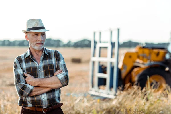 Self-employed farmer in straw hat standing with crossed arms near tractor — Stock Photo