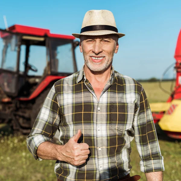 Fröhlicher Bauer mit Strohhut zeigt Daumen hoch neben Traktor — Stockfoto