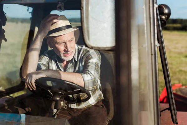 Selective focus of senior farmer touching straw hat while driving tractor — Stock Photo