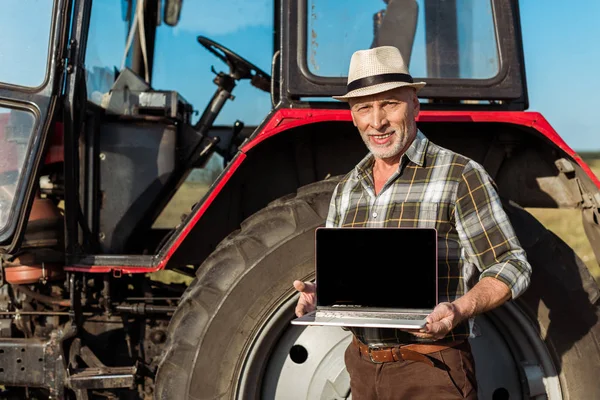 Granjero feliz en sombrero de paja que sostiene el ordenador portátil con pantalla en blanco cerca del tractor - foto de stock