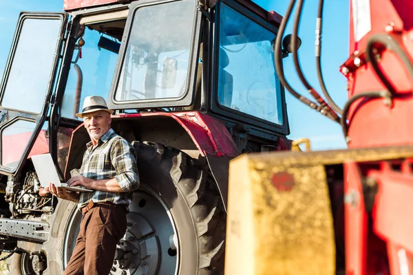 Messa a fuoco selettiva di felice agricoltore in cappello di paglia utilizzando computer portatile vicino trattore — Foto stock