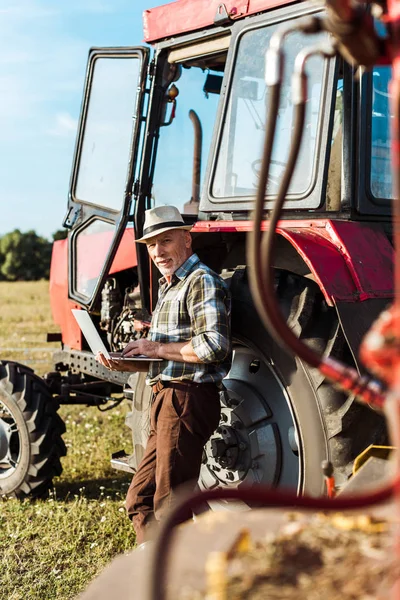Selective focus of farmer in straw hat using laptop near tractor — Stock Photo