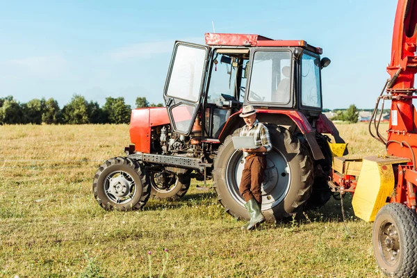 Contadino barbuto in cappello di paglia utilizzando computer portatile vicino al trattore — Foto stock