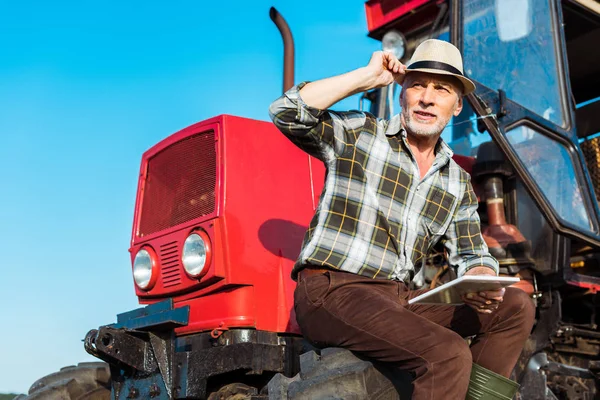 Senior farmer in straw hat holding digital tablet near red tractor — Stock Photo