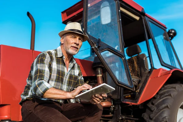 Agricultor senior en sombrero de paja utilizando tableta digital cerca de tractor rojo - foto de stock