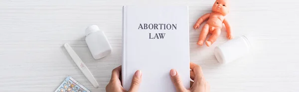 Foto panorámica de la mujer sosteniendo libro con letras de aborto cerca de muñeca y píldoras del aborto - foto de stock