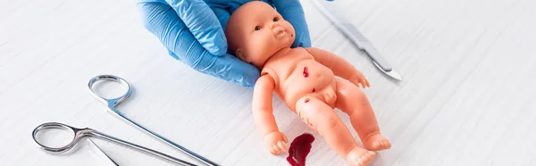 Panoramic shot of doctor in blue latex gloves holding baby doll with blood near medical instruments — Stock Photo