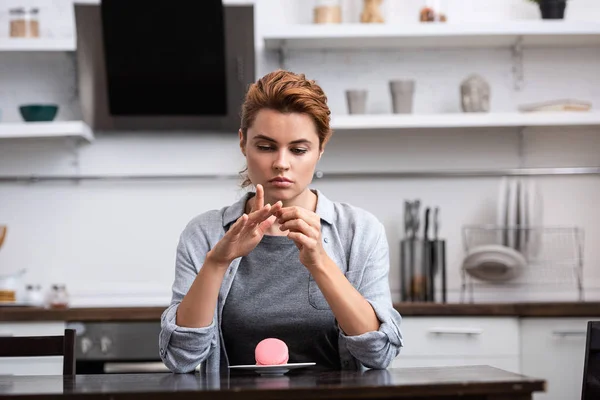 Attraktive Frau, die Hände betrachtet, während sie neben Untertasse mit rosa Dessert sitzt — Stockfoto