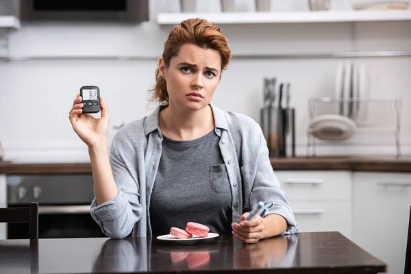 Mujer molesta con alergia dulce sentado cerca de postre rosa y la celebración de monitor de glucosa - foto de stock