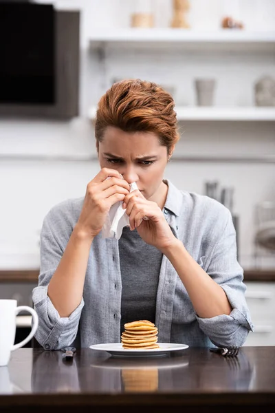 Sick woman sneezing in napkin near pancakes at home — Stock Photo