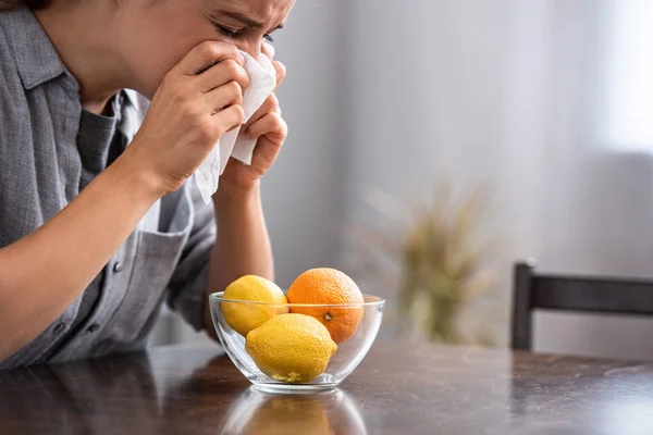 Woman with closed eyes sneezing in tissue near orange and lemons in bowl — Stock Photo