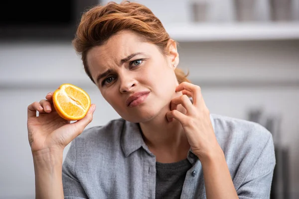 Upset woman holding half of orange while scratching neck — Stock Photo
