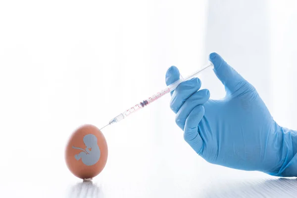 Cropped view of doctor in latex glove holding syringe near egg with baby silhouette — Stock Photo