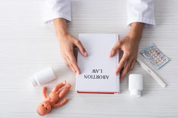 Top view of woman holding book with abortion letters near baby doll and abortion pills — стоковое фото