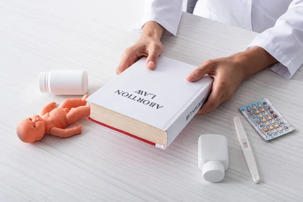 Cropped view of doctor holding book with abortion lettering near baby doll and abortion pills — Stock Photo