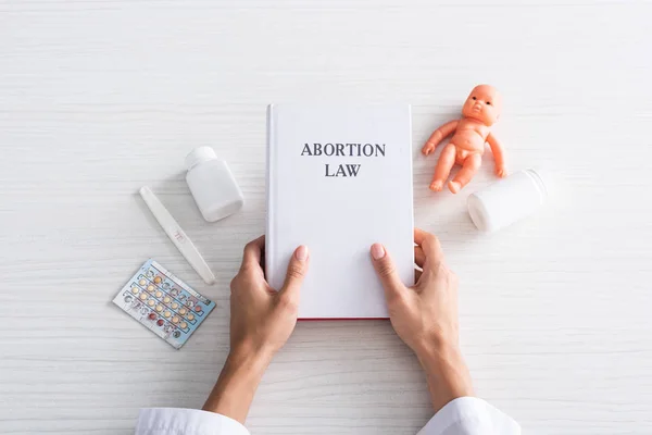 Top view of woman holding book with abortion lettering near baby doll and abortion pills — стоковое фото
