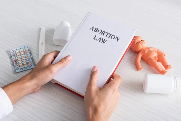 Cropped view of woman holding book with abortion lettering near baby doll and abortion pills — Stock Photo