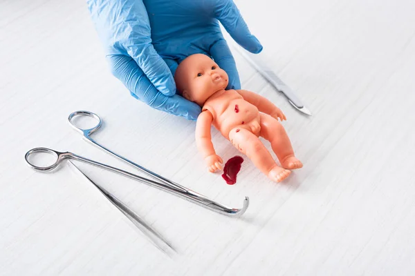 Cropped view of doctor in blue latex gloves holding baby doll with blood near medical instruments — Stock Photo