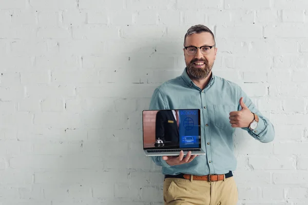 Guapo y sonriente hombre de negocios mostrando el pulgar hacia arriba y la celebración de la computadora portátil con el sitio web de reserva - foto de stock