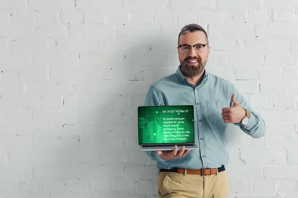 Guapo y sonriente hombre de negocios mostrando el pulgar hacia arriba y la celebración de la computadora portátil con el sitio web de salud - foto de stock